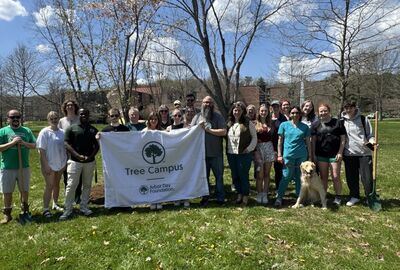 A diverse group of smiling people stand on grass in a park, holding a "Tree Campus" banner. A golden retriever sits beside them.