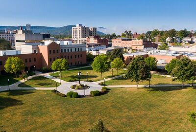 Aerial view of a college campus with green lawns, brick buildings, and pathways. Trees and a circular garden, including a clock tower, dot the scene