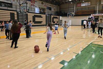 Students engage in basketball activities in a gymnasium. The scene is lively, with energy as students dribble and socialize under a bright, organized setting.