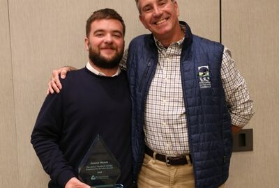 Two men pose smiling, one holding a triangular award. They appear happy and proud. One wears a plaid shirt with a blue vest, the other a dark sweater.