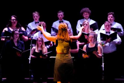 A choir conductor leads a group of men and women singing on stage. The conductor's arms are raised, and the singers hold sheets, creating a harmonious scene.