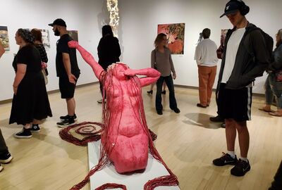 In a gallery, visitors explore art. A central pink sculpture of a reclining figure with outstretched arms and cascading hair catches attention.