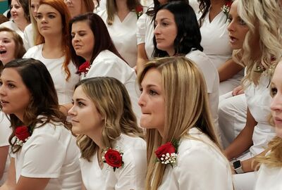 A group of women in white uniforms with red rose corsages sit closely together at a ceremony.