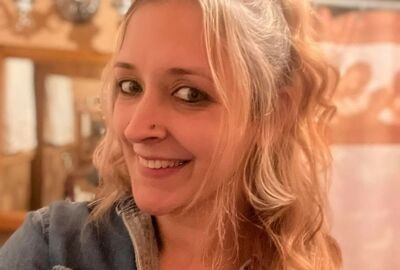 Smiling woman with wavy blonde hair in a casual denim jacket stands in a cozy room with soft lighting and subtle background decor.