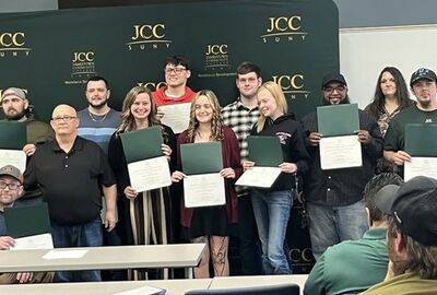 A group of people stand proudly holding certificates in front of a JCC SUNY backdrop. Smiles convey a sense of achievement and celebration.