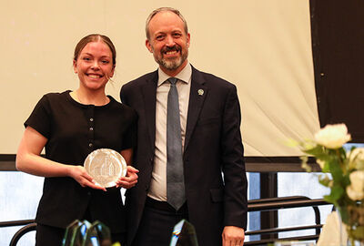 A smiling woman in a black dress holds an award next to a man in a suit. They're indoors with a light background.