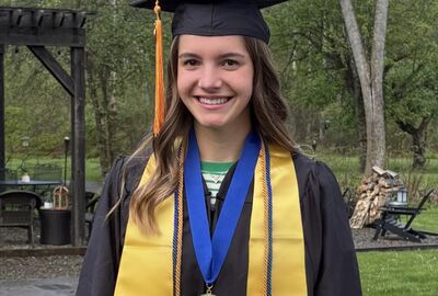 A young woman wearing graduation regalia stands outdoors smiling at the camera. She is dressed in a black cap and gown with gold honor stoles, blue and gold cords, and a medal on a blue ribbon around her neck. Trees and a patio area are visible in the background.