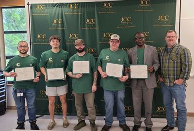 Six people stand smiling in front of a "JCC Workforce Development" banner. Five hold certificates, wearing green shirts, conveying a celebratory mood.