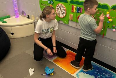 A young boy interacts with a colorful sensory wall panel in a brightly lit room, while an adult sits nearby observing and holding a stylus.