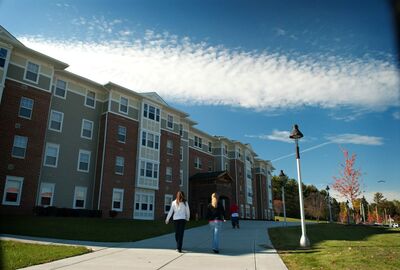 Exterior of a multi-story dormitory building with brick and siding, viewed from a sidewalk on a clear day with scattered clouds.