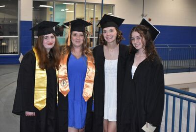 Four graduates in black caps and gowns pose happily. Two wear gold and orange stoles. They stand indoors, smiling, conveying a sense of achievement.