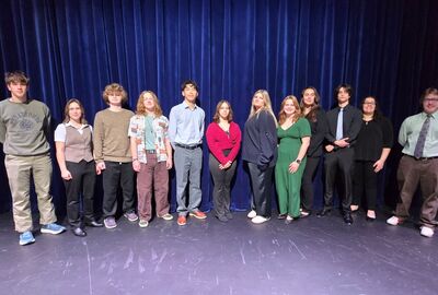 A diverse group of young people stands in a line on a stage in front of a dark blue curtain.