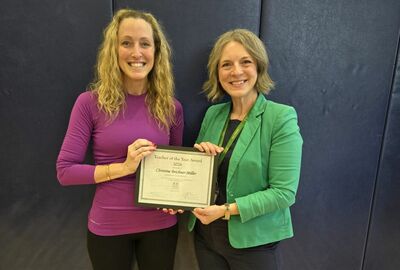 Two women stand together, one on the left wearing a purple long-sleeved shirt and black leggings, and the other on the right wearing a green blazer over a dark top. They are both smiling and holding a framed "Teacher of the Year Award" certificate dated 2026.