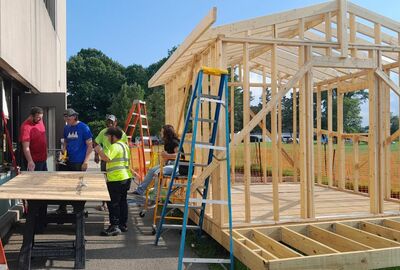 Construction technology students collaborating on a wooden structure, using ladders and tools in a sunny outdoor setting.