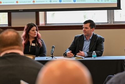 Two individuals sit at a table on a panel at an event, with one speaking and gesturing with their hands.
