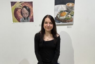 A person with dark hair smiles while sitting in front of two paintings on a white wall, one painting depicting a stylized portrait with a distressed expression, and the other a still life of avocado toast and coffee.
