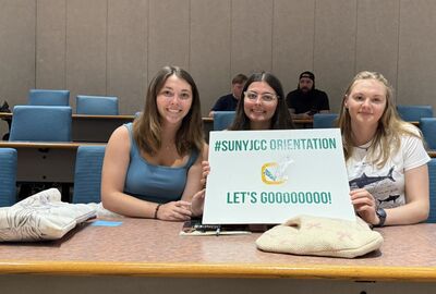Three women seated in a college classroom, holding a sign that reads #SUNYJCC orientation, let's gooooooo!