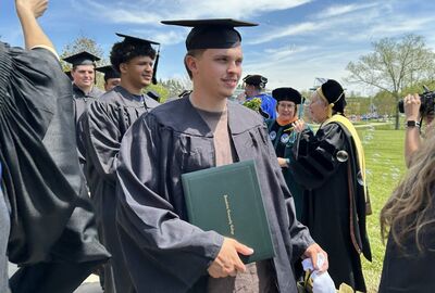Graduates in academic regalia walk across a grassy field under a partly cloudy sky. A graduate in the foreground holds a green diploma cover.