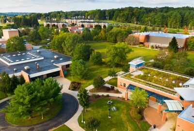 Aerial view of a university campus with brick buildings, green spaces, and mature trees under a cloudy sky. One building features a green, vegetated roof and blue architectural accents.