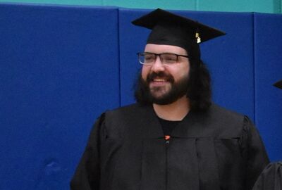 A smiling man with long dark hair, glasses, and a beard wears a black graduation cap and gown. He is standing in front of a blue wall.
