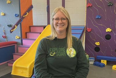A smiling blonde woman wearing glasses and a dark green long-sleeved shirt stands in a colorful indoor play area. The background features a yellow slide, pink stairs, and a purple climbing wall.