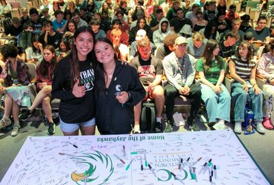 Two smiling young women stand in front of a large banner covered in signatures and a "Home of the Jayhawks" logo, with a seated audience behind them.