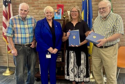 A group of four people stand together, holding two blue books. An American flag and two other flags are visible behind them.