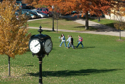 A black double-sided park clock stands in the foreground with a group of people walking across a grassy quad in the background, surrounded by trees with autumn foliage.