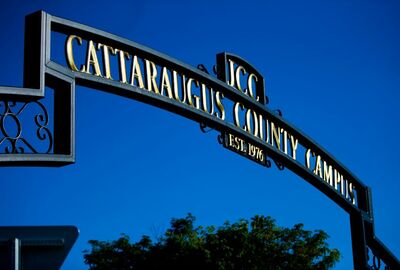 Ornate black metal archway reading “Cattaraugus County Campus” against a vivid blue sky, with trees below; the grand entrance feels formal and welcoming.