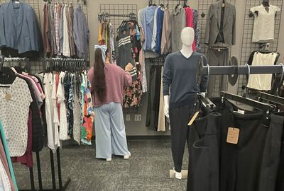A person in a pink sweater and light blue wide-leg pants browses clothing racks in a retail store, with a male mannequin dressed in a blue sweater and dark pants displayed prominently.
