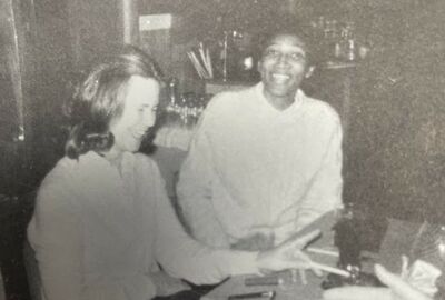 Two individuals sit at a table in a dimly lit indoor setting, with one person in the foreground laughing and holding thin sticks, and the other person in the background smiling.