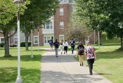 A group of students walk away from the camera on a paved path through a grassy campus with trees and a brick dormitory building in the background.