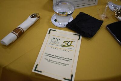 A gold fork and spoon wrapped in a white napkin with brown stripes sits on a yellow tablecloth next to a program for the Jamestown Community College Cattaraugus County Campus 50th Anniversary Celebration on Thursday, September 10, 2026.
