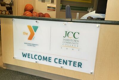 Clear glass sign on a reception desk with the YMCA logo and "Jamestown Community College YMCA" on the left, and the "JCC Jamestown Community College SUNY" logo on the right, with "WELCOME CENTER" printed in large teal letters below. Several basketballs are visible on the counter behind the sign.