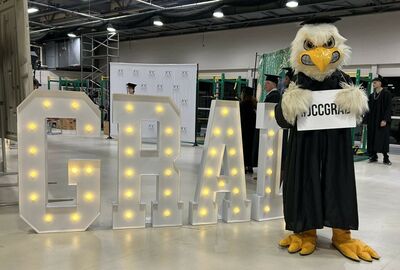 Costumed eagle mascot in a black robe stands beside large glowing “GBA” marquee letters in an industrial event space, creating a festive mood.
