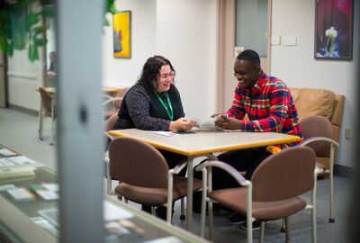 Two smiling adults sit across a table in a bright office, sharing a friendly conversation; their relaxed posture creates a warm, welcoming tone.