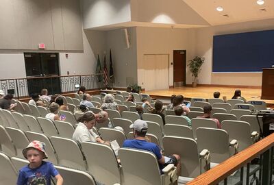 An audience of people sit in tiered seating facing a wooden stage in an auditorium.