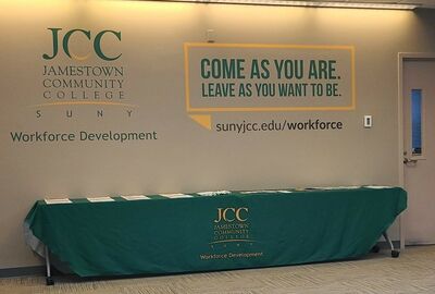 Jamestown Community College logo and "Workforce Development" text on a wall, with a banner displaying "Come as you are. Leave as you want to be." and the website sunyjcc.edu/workforce. A green tablecloth-covered table with brochures is in the foreground.
