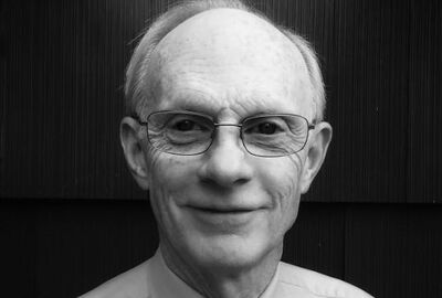 Black-and-white close-up portrait of an elderly bald man wearing glasses, smiling gently against a dark wooden background; the mood is warm and thoughtful.