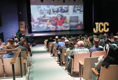 An audience fills a theater-style hall, watching a presentation featuring a woman on screen as illuminated JCC letters glow nearby.