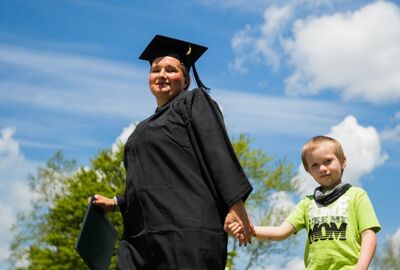 A proud graduate in a black cap and gown holds a diploma folder while walking hand in hand with a young boy beneath a bright blue sky.